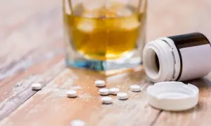 Szklanka z bursztynowym płynem i rozsypane tabletki obok otwartego pojemnika. Uważaj na interakcje antybiotyk a alkohol.