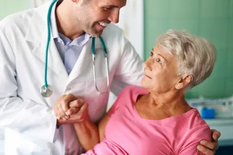 Lekarz w białym kitlu wspiera starszą pacjentkę. To przykład troski w geriatrii, która zajmuje się zdrowiem seniorów.
