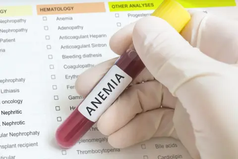 Ręka w rękawiczce trzyma probówkę z krwią z napisem "ANEMIA". Obok znajduje się lista badań, gdzie zaznaczono "Anemia", sugerując diagnostykę i poszukiwanie sposobów, jak leczyć anemię.